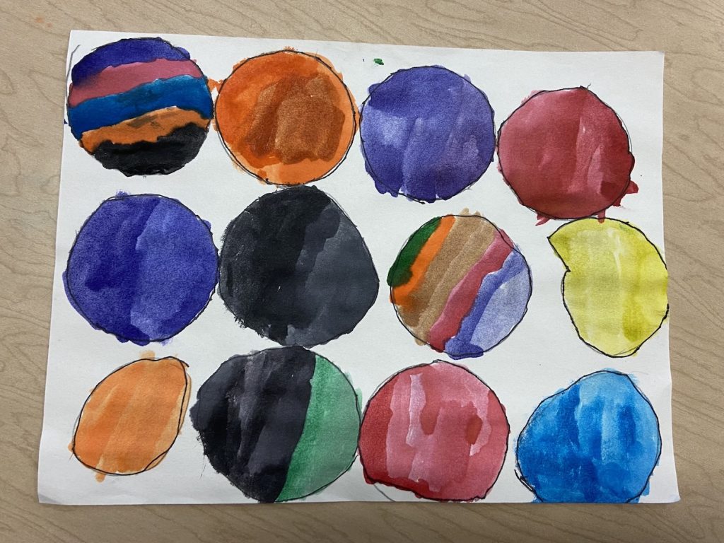 A student’s watercolor art piece features twelve hand-drawn circles, each filled with different colors and patterns. The circles are arranged in a grid on white paper and showcase a range of blending techniques, from solid fills to layered and striped effects.